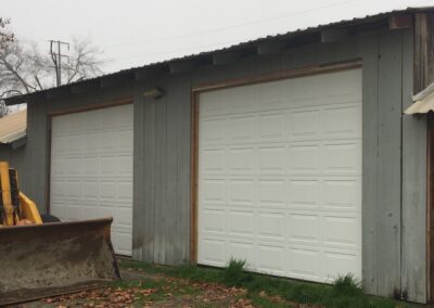 Two white garage doors on a gray wooden structure with a yellow bulldozer's scoop visible on the left.